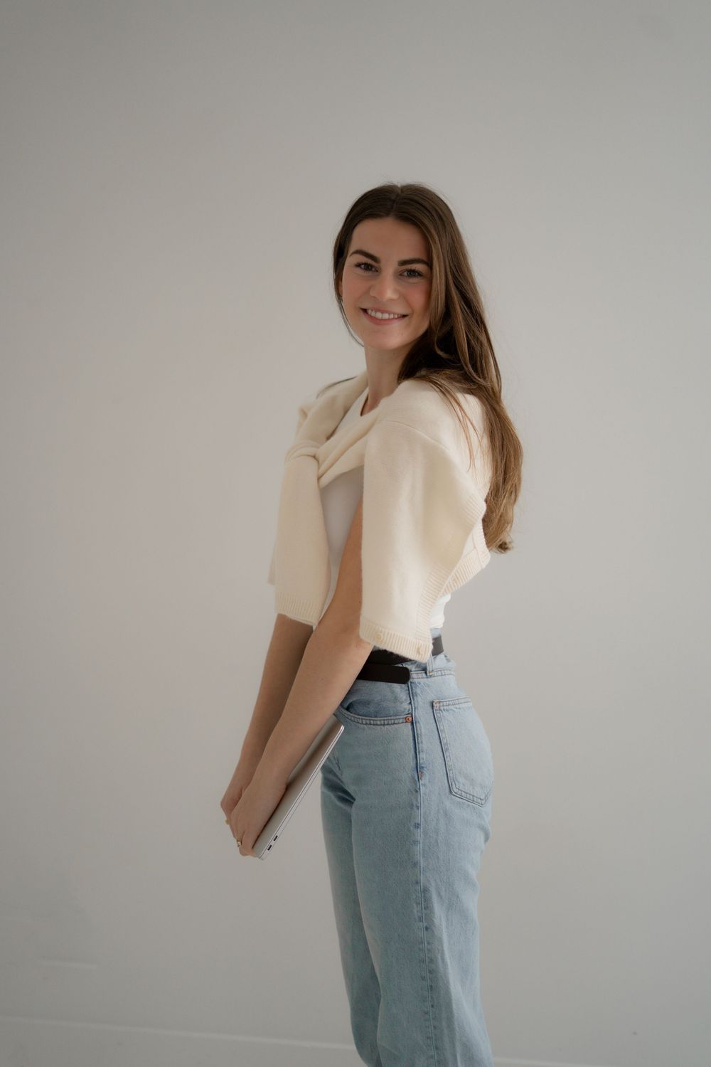 A person in cream top and light jeans poses naturally against white wall.