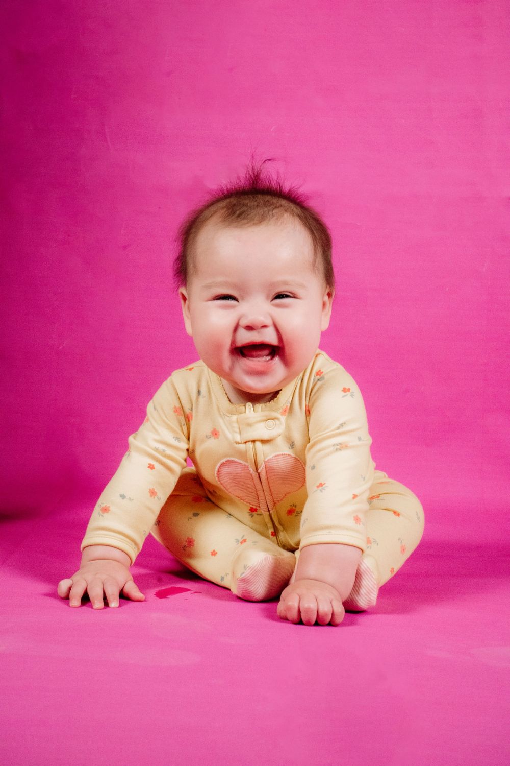 Laughing baby in yellow onesie sitting on pink background.
