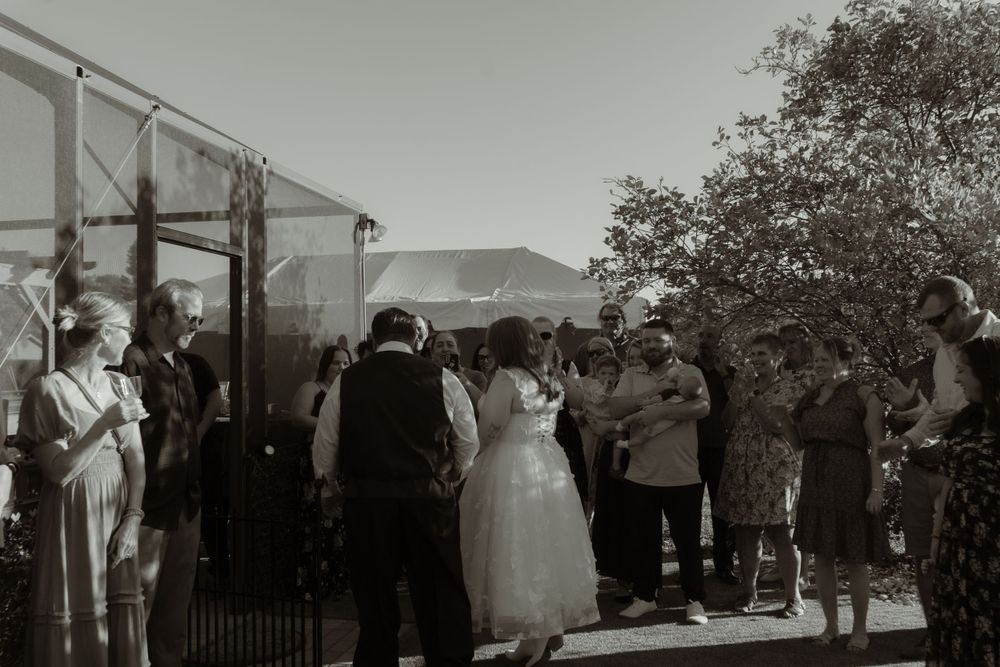Black and white photo of guests gathered around the bride and groom during a candid moment at an outdoor wedding reception.