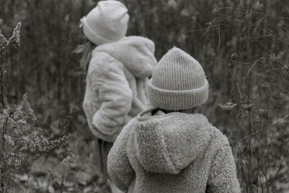A black and white image of two children in winter attire walking through tall grass.
