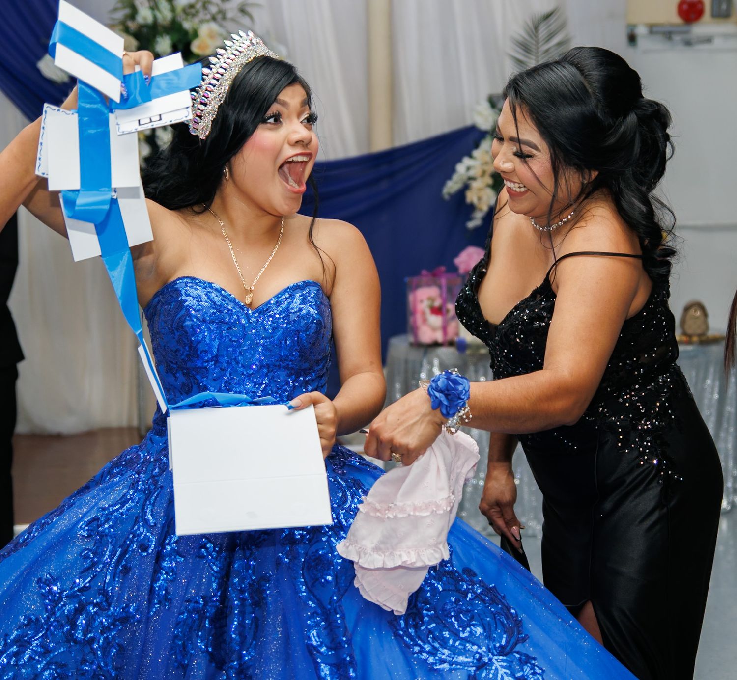 Celebratory moment at a quinceañera as two people share laughter while opening presents in blue and black dresses.