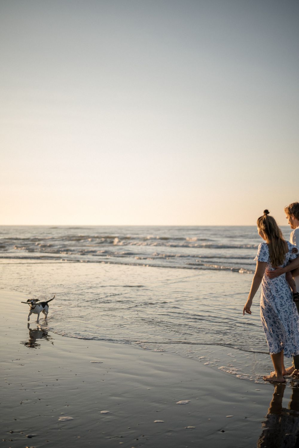 A dog plays in the waves as someone in a floral dress walks along the beach at sunset.