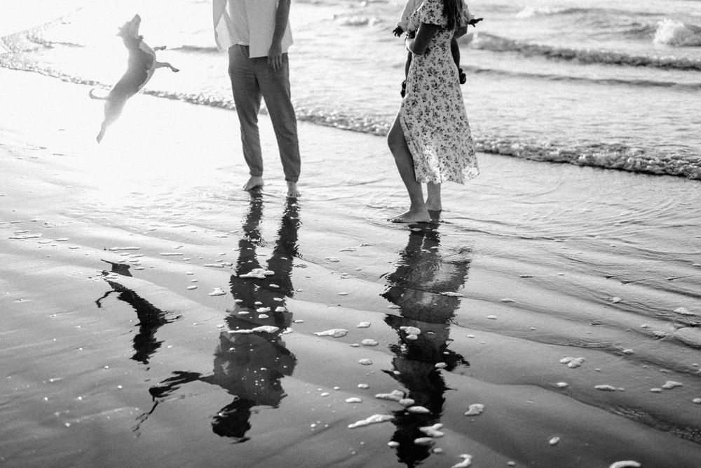 Black and white photo of people's reflections in wet sand at the beach.