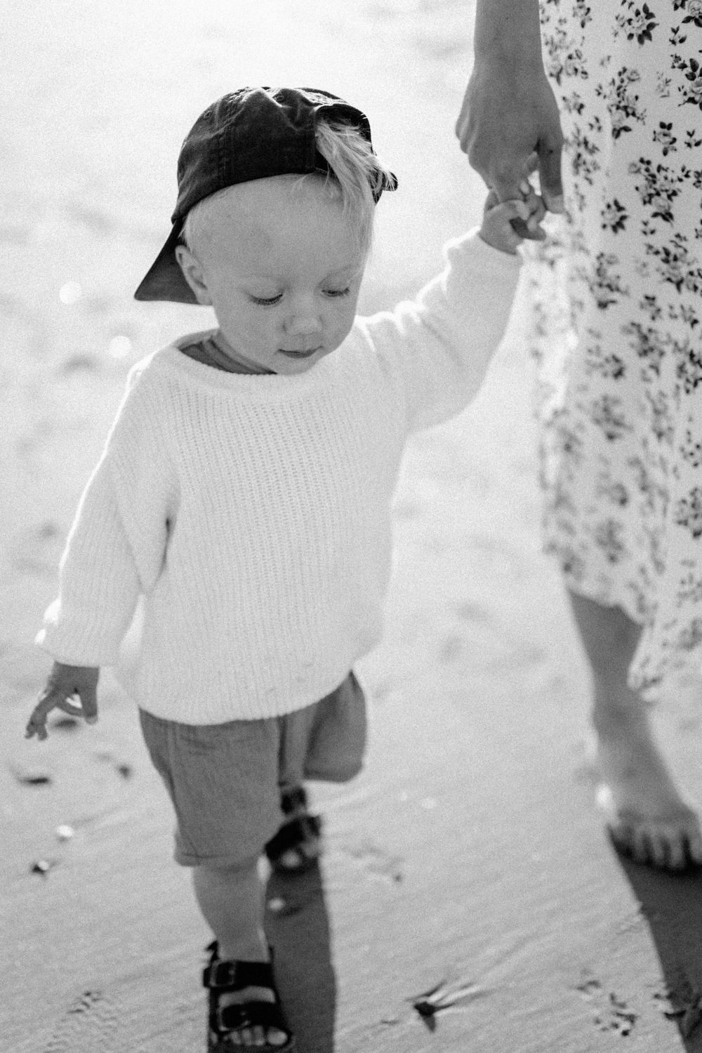 A toddler in a white sweater and cap walks on the beach holding someone's hand.
