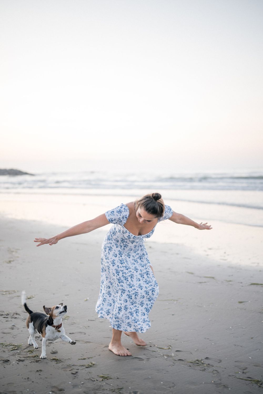 A person in a floral dress plays with a small dog on the beach at sunset.