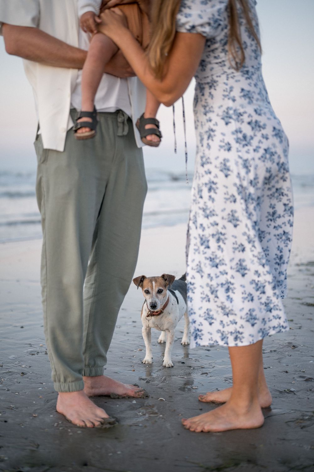 Bare feet in sand with small dog standing between people wearing casual beach attire.