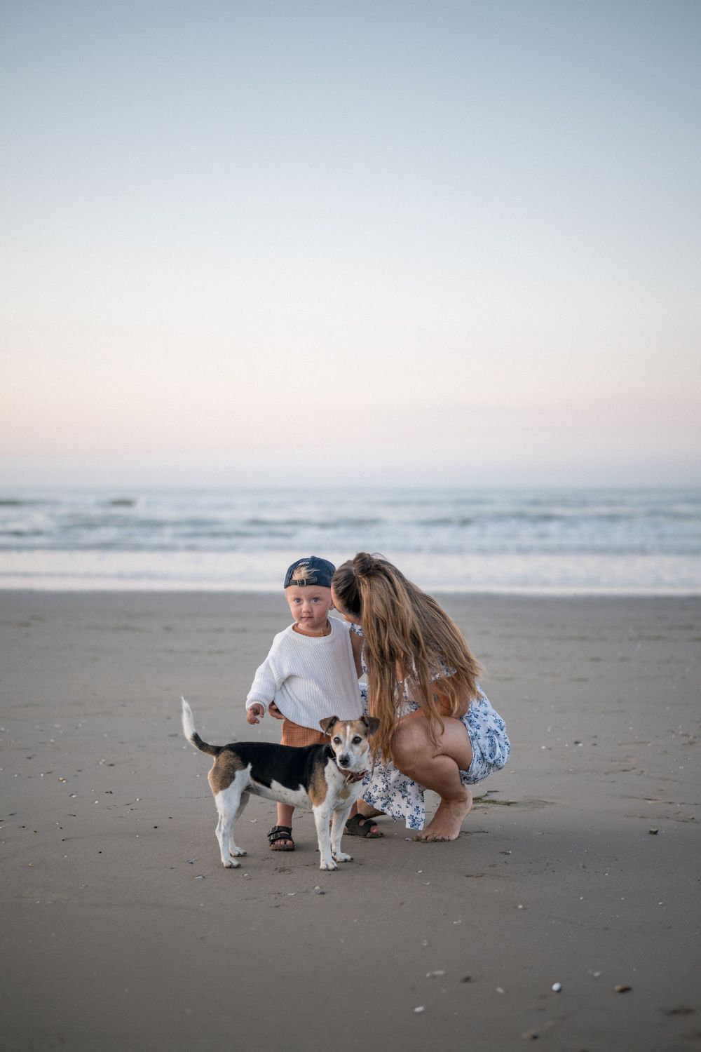 Mother and child petting a small dog on a serene beach at dusk.
