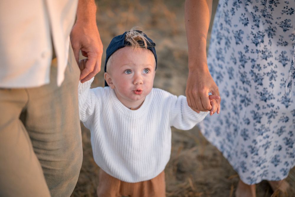 Young child in a white sweater holding hands with adults on a beach.