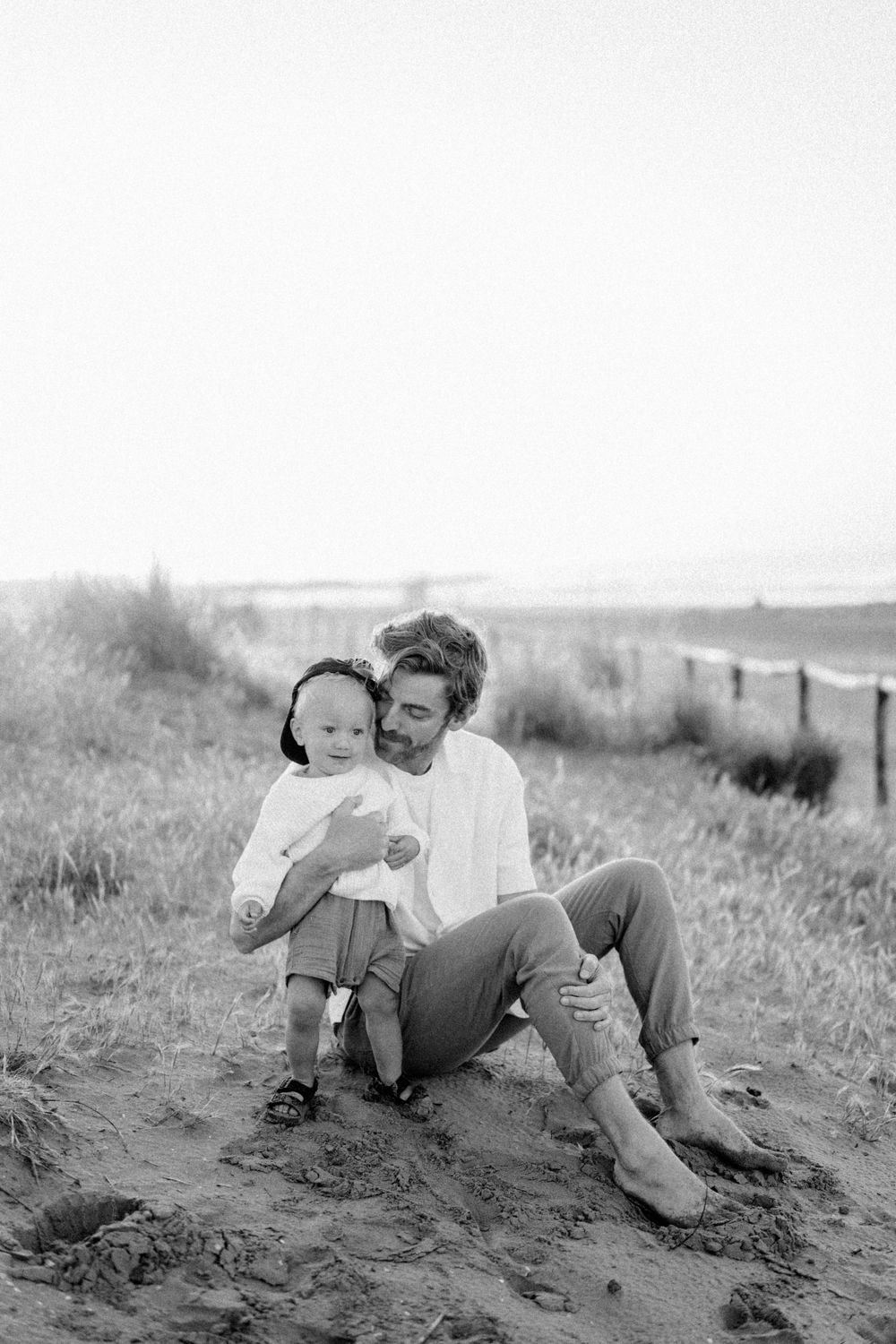 A tender black and white portrait of a parent and child sitting together on sandy ground near the beach.