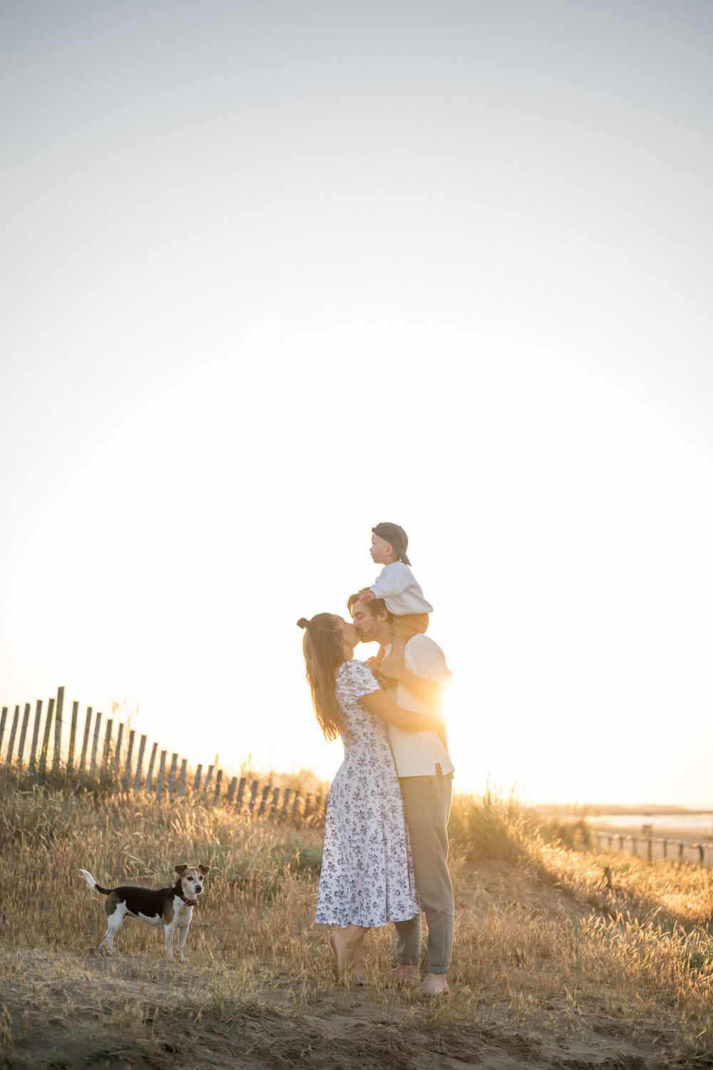 A silhouetted family lifts their child against a golden sunset with their dog nearby.