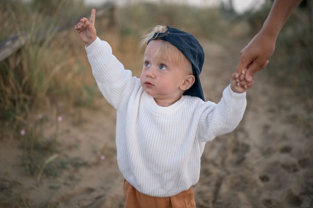 Small child in white sweater and blue hat pointing upward on beach path.