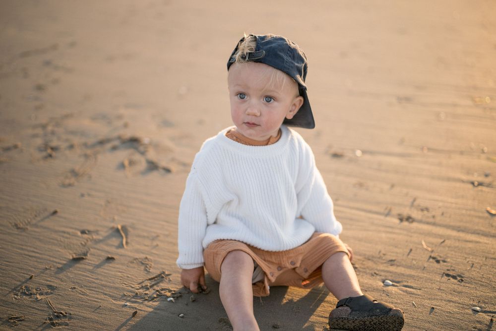 Toddler in baseball cap and white sweater sitting on sandy beach.