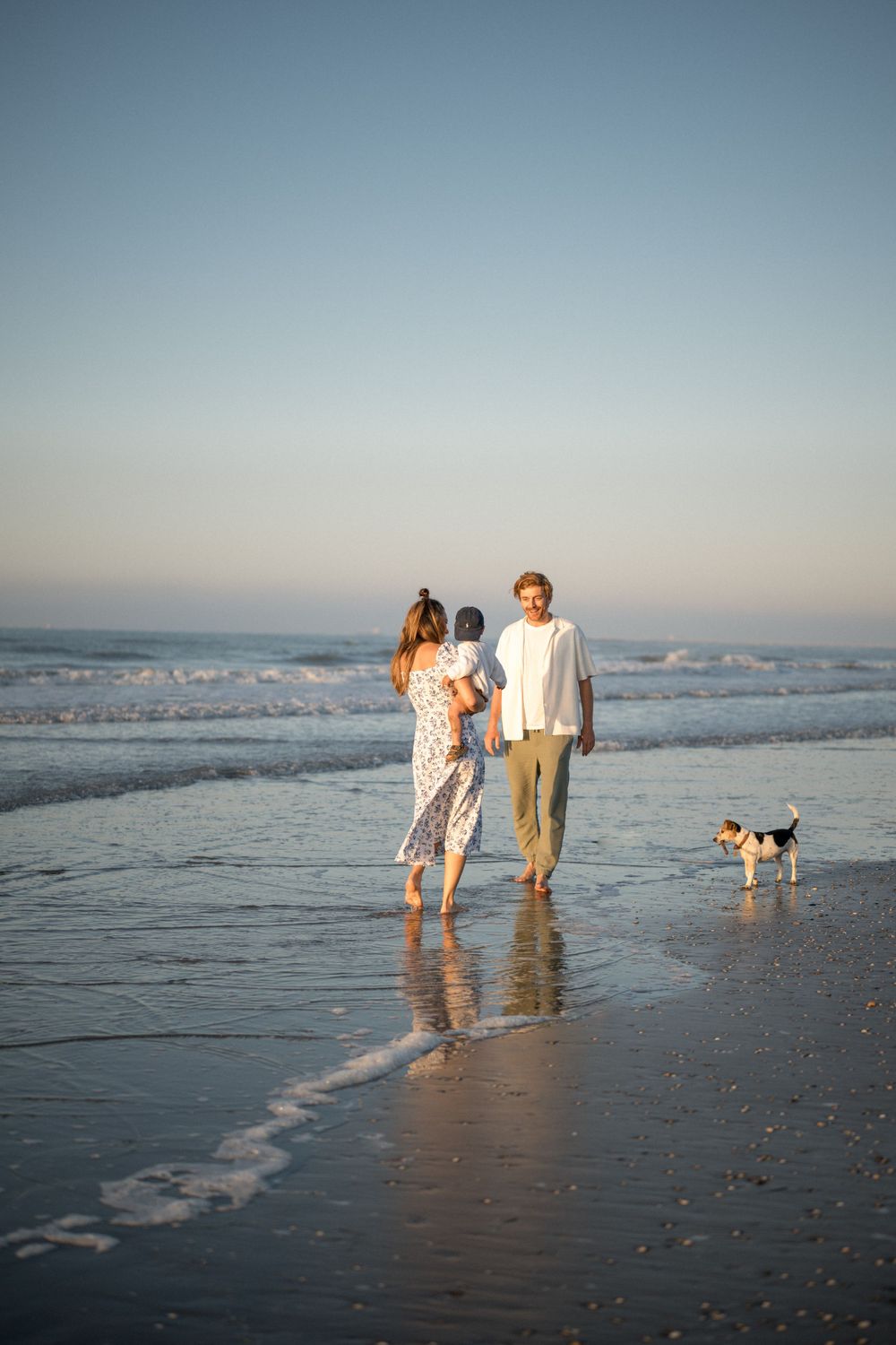 A couple walks along the beach at sunset with their small dog following in the surf.