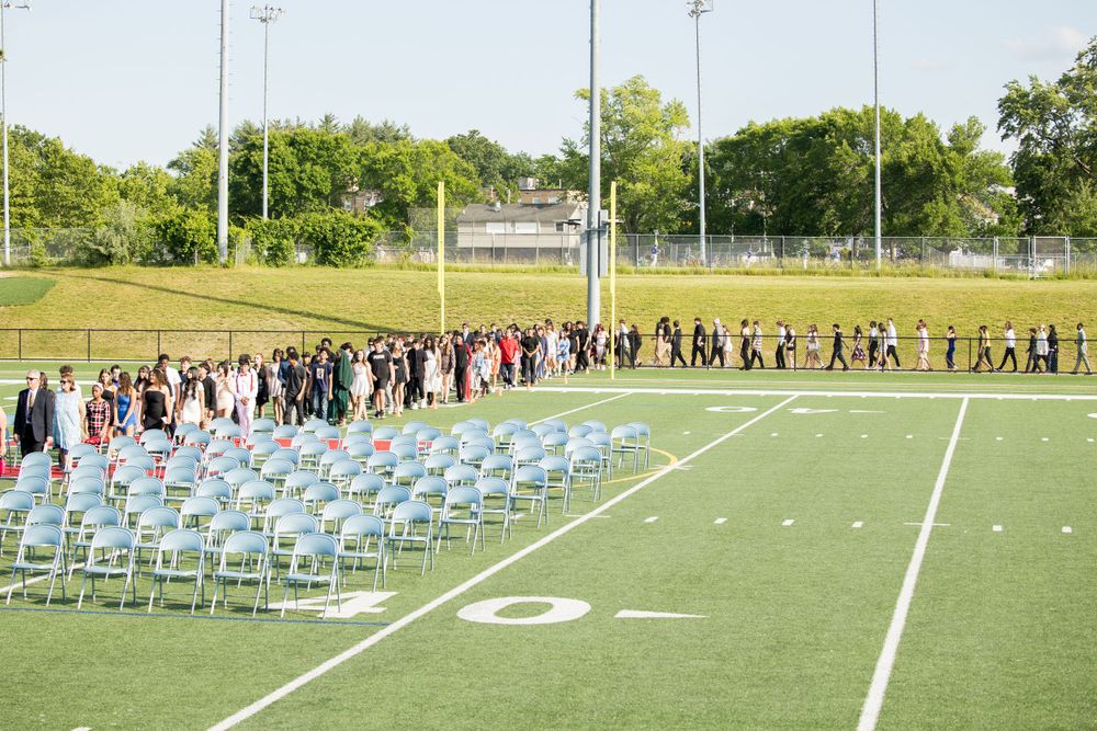 A line of graduates in formal attire walks across a football field with rows of empty chairs set up for a ceremony.