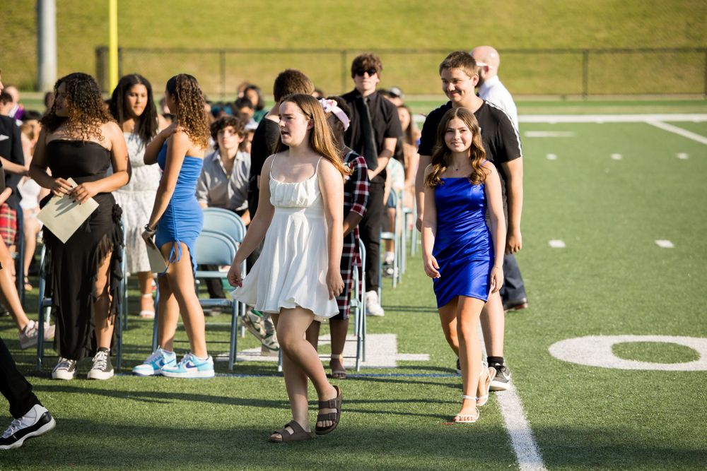 Students in formal dresses and suits walk along the field during what appears to be a graduation ceremony.