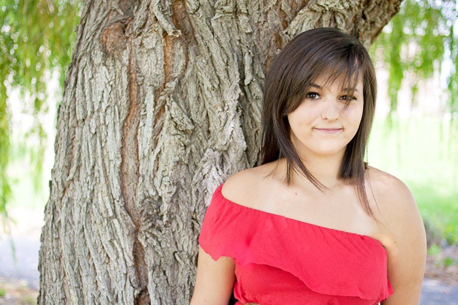 Someone wearing a red off-shoulder dress poses casually against a textured tree trunk.