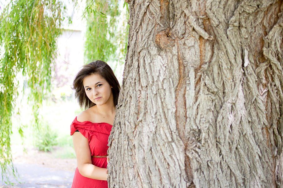 A portrait featuring someone in a red dress peeking from behind a large tree trunk.