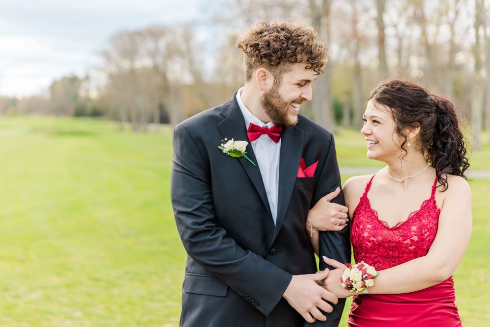 Formal couple in red dress and black tuxedo smiling together on grassy lawn.