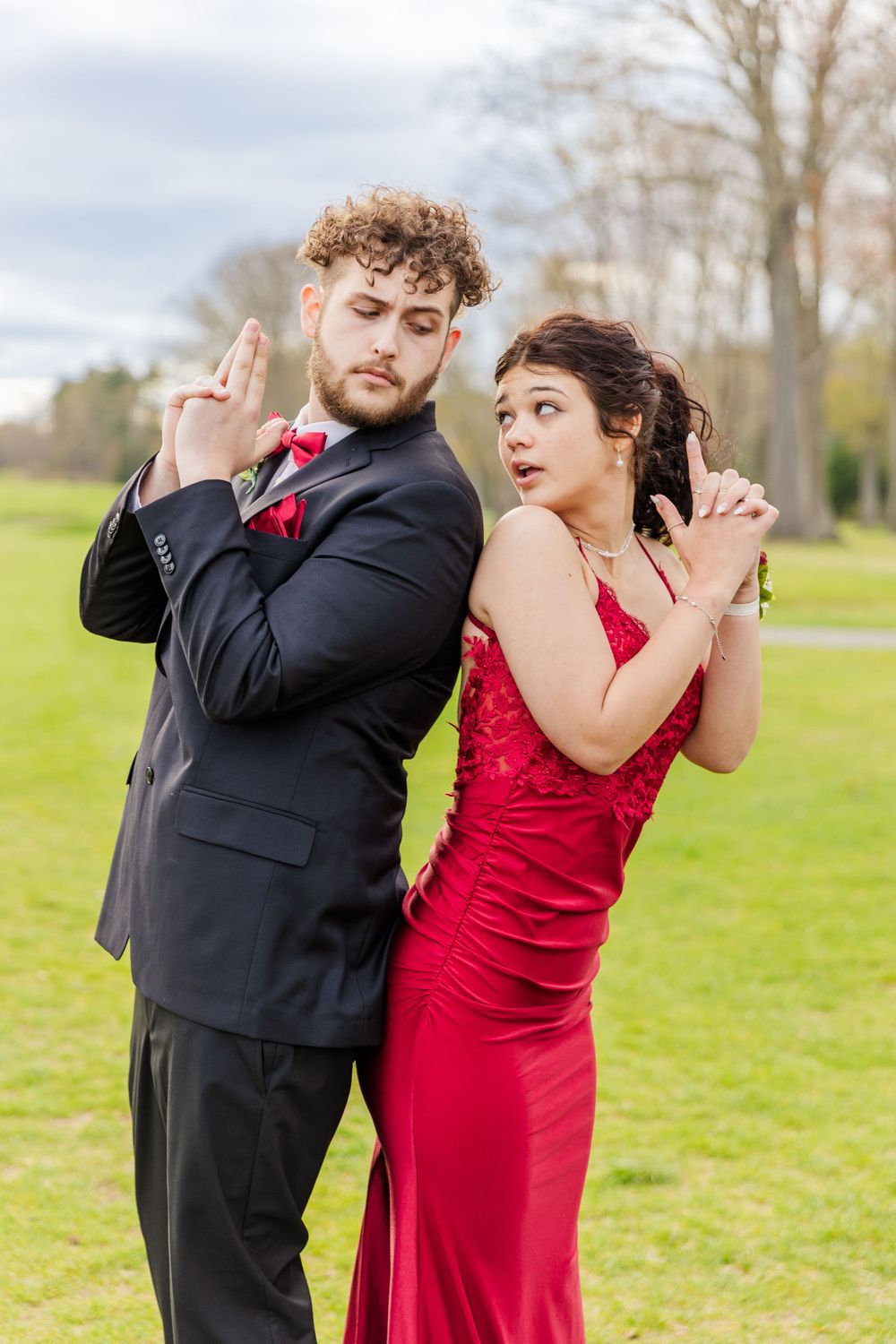 Couple in formal attire striking playful pose with finger guns on green lawn.