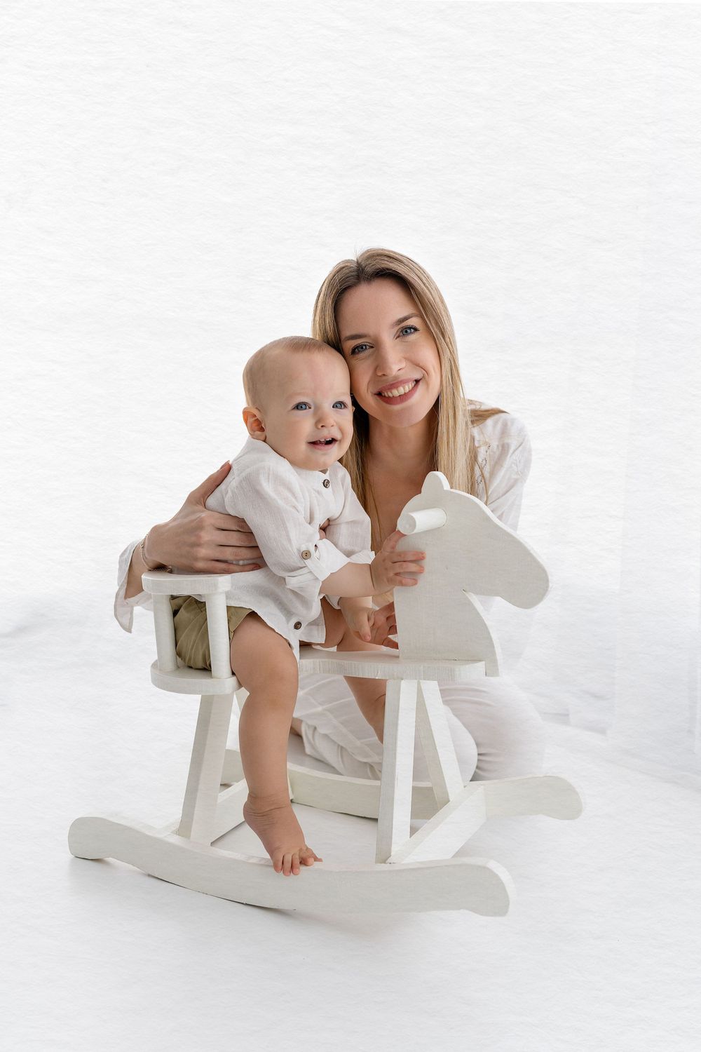 Baby and adult share happy moment on white rocking horse.