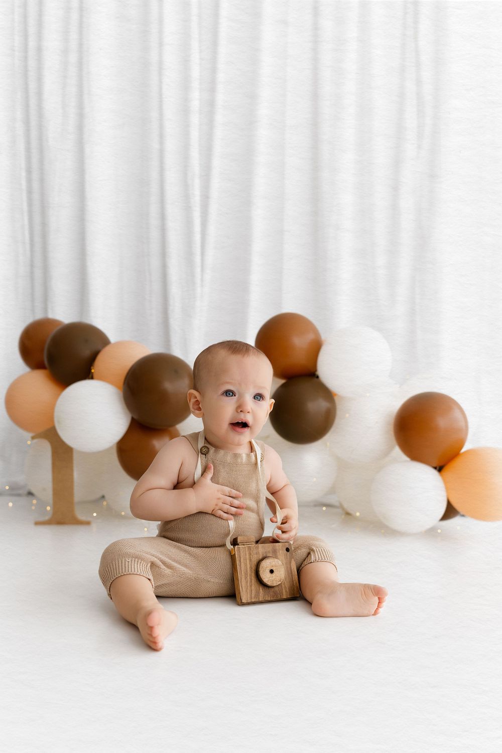 Baby sitting in beige outfit with brown and white balloon garland background.