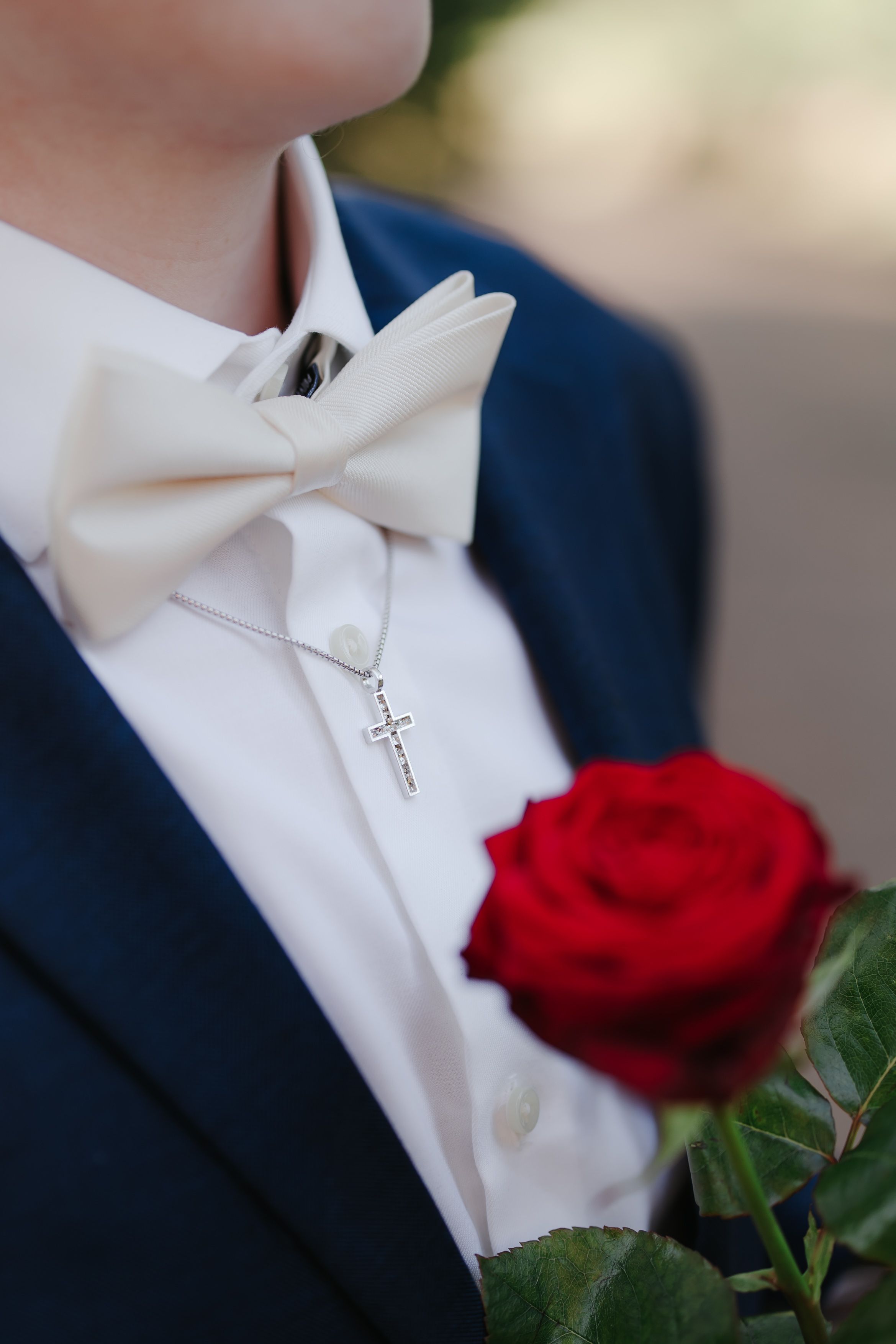 Man in navy suit with white bow tie, cross necklace, holding a red rose, formal attire for wedding or prom.