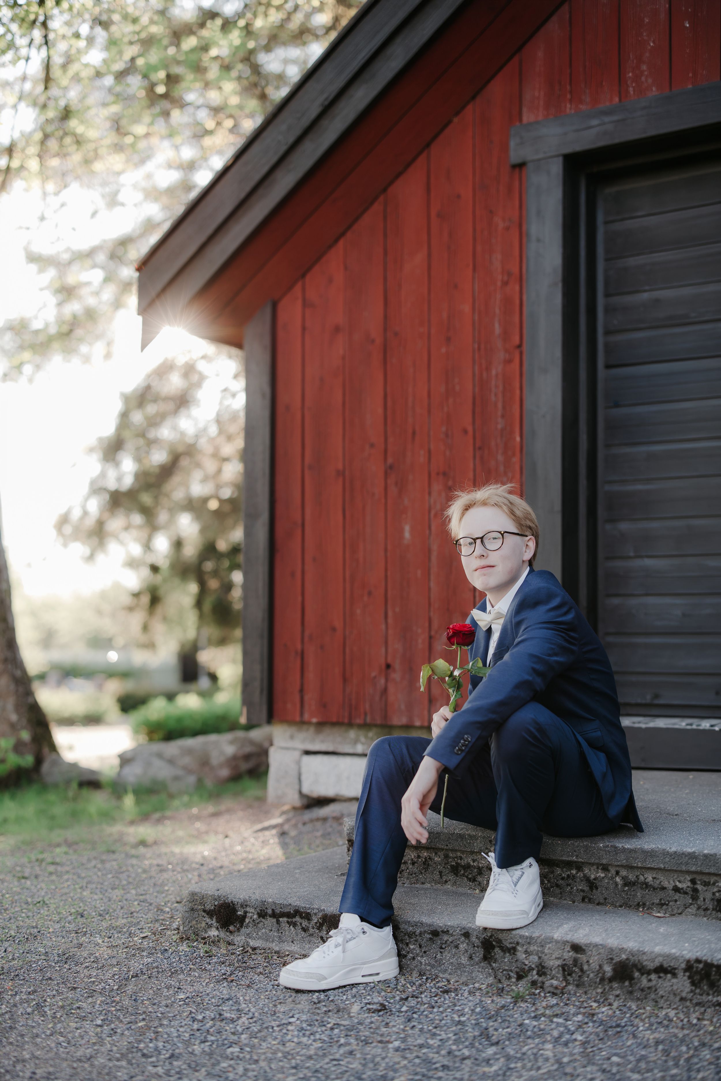 Young boy in blue suit holding red rose sits on steps of rustic red wooden building outdoors.