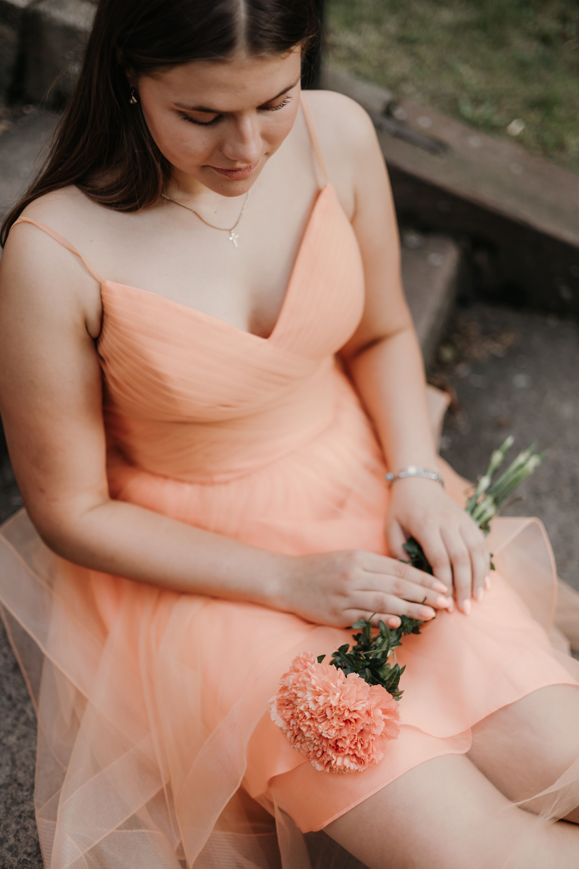 Person in peach-colored dress holding coral carnations while seated outdoors.