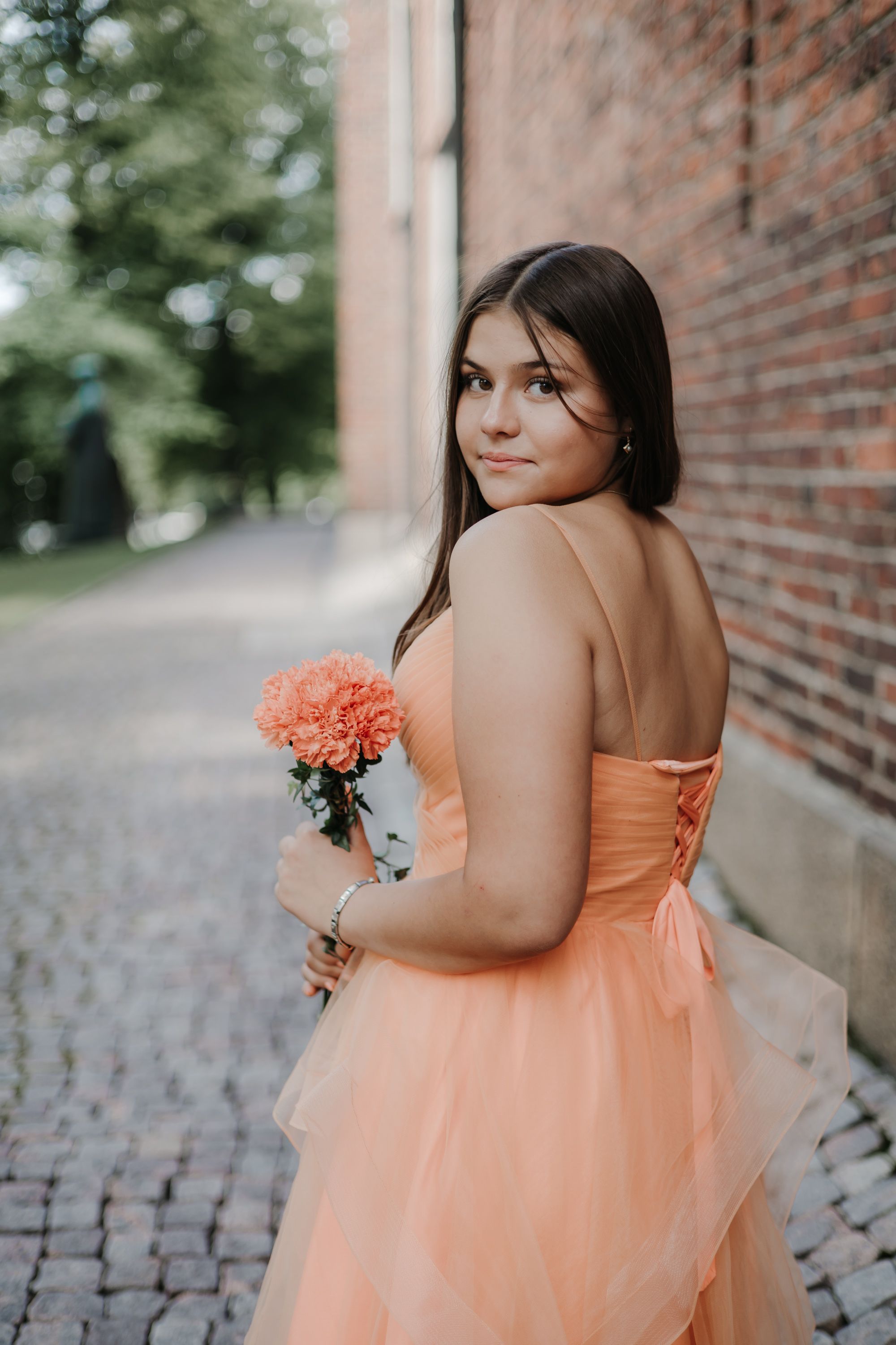 Bridesmaid in flowing peach gown holds bouquet while posing near brick wall.
