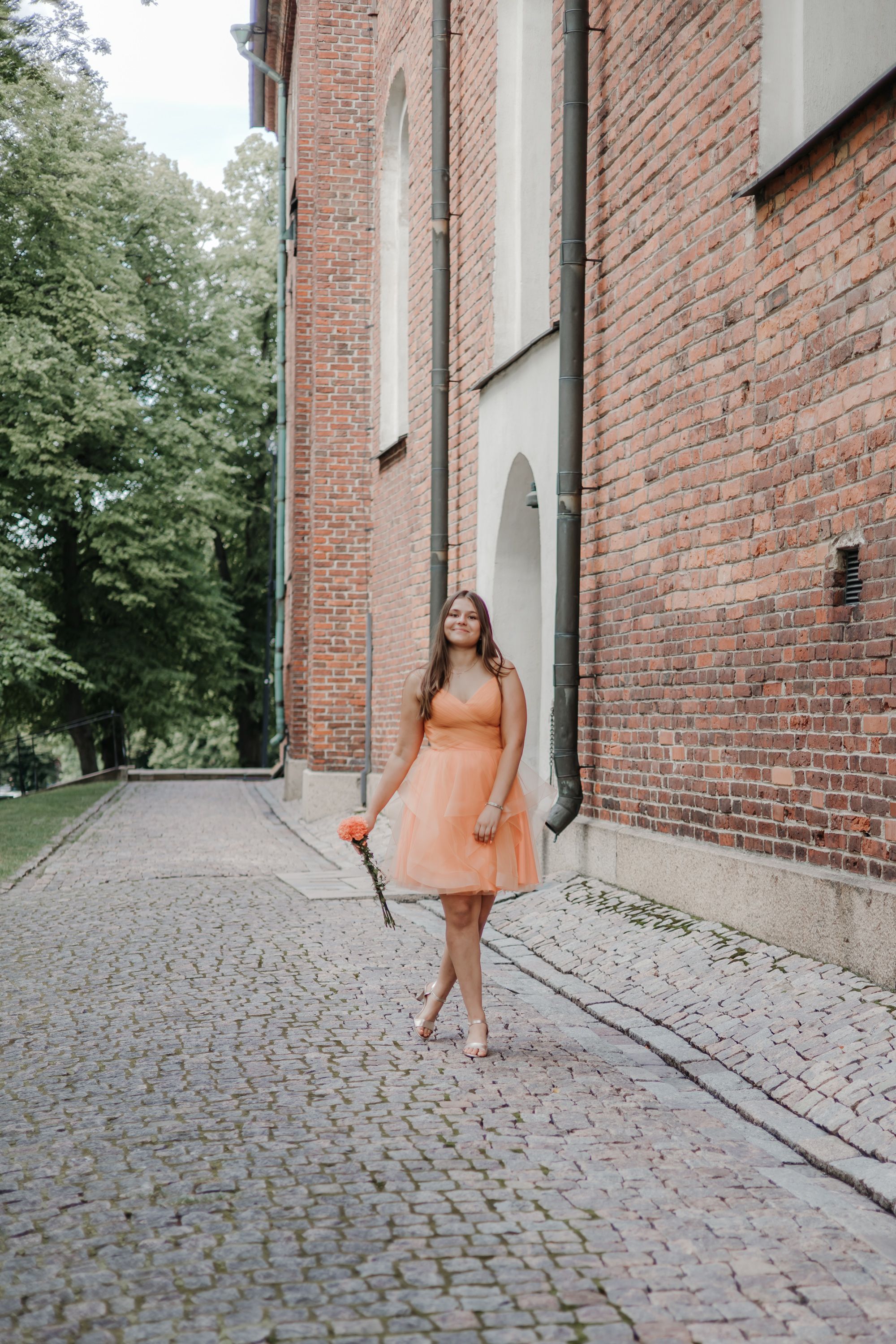 A person in a peach-colored dress poses against a brick building wall on a cobblestone path.