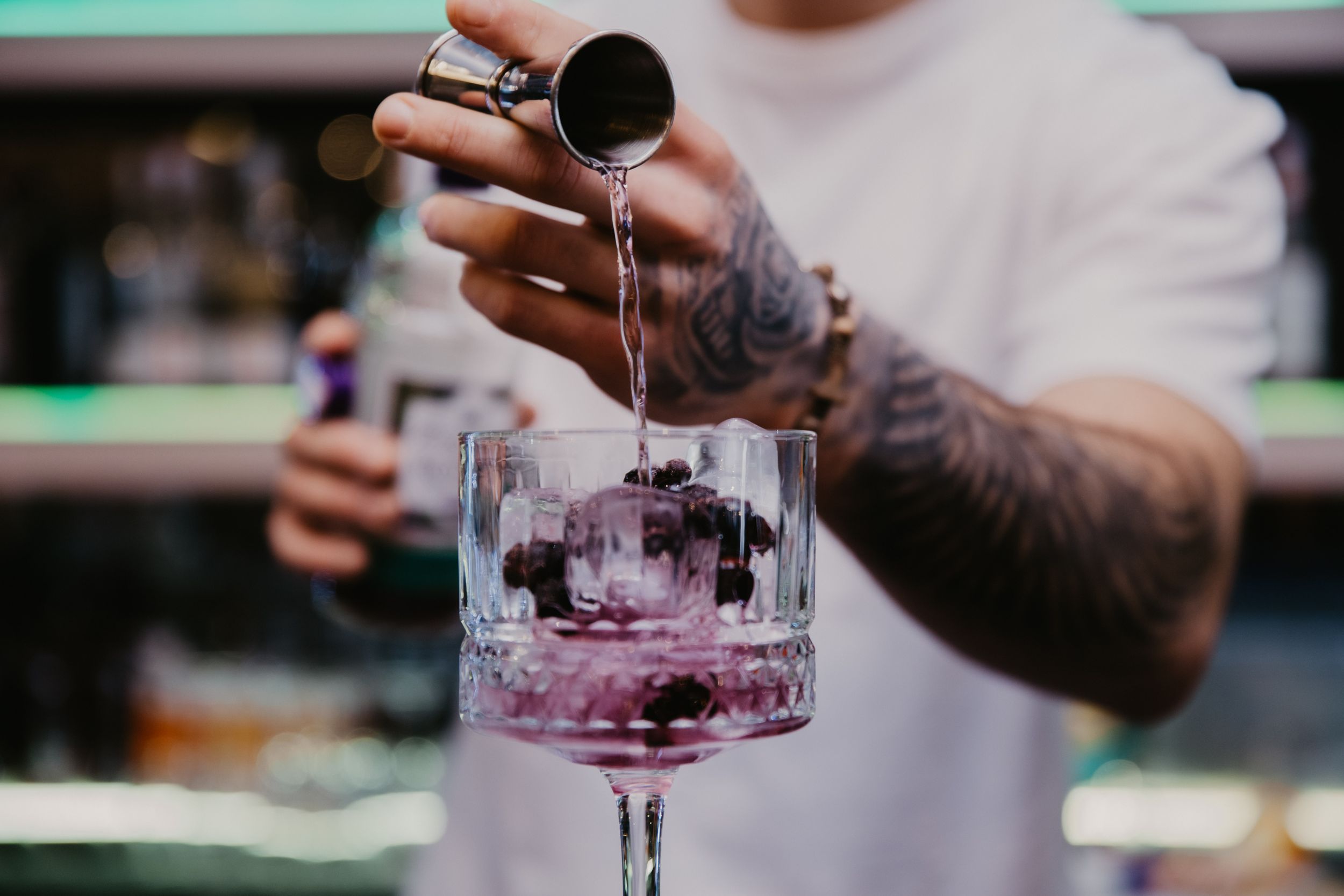 Tattooed hands pouring purple liquid into crystal cocktail glass.