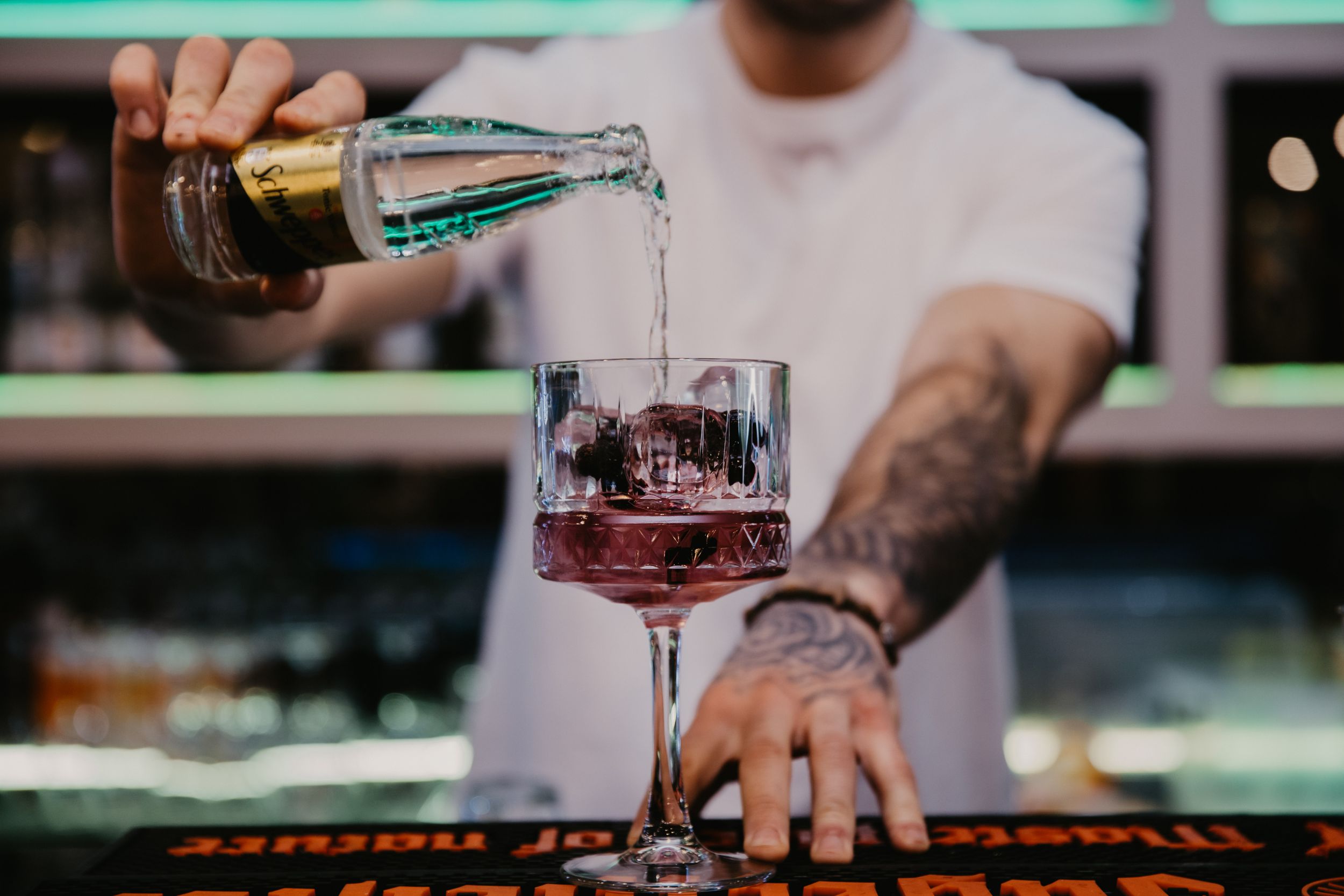 Bartender pouring a measured drink into a wine glass from a bottle at a bar counter.