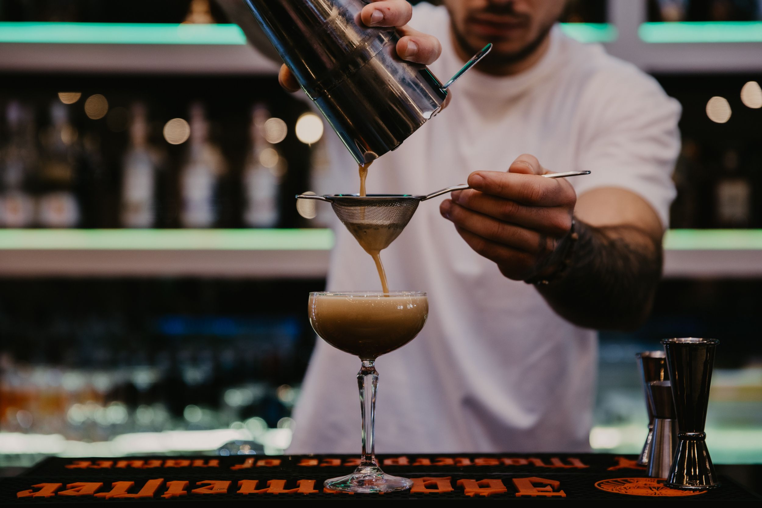 Cocktail being carefully poured from a shaker into a martini glass at a bar.