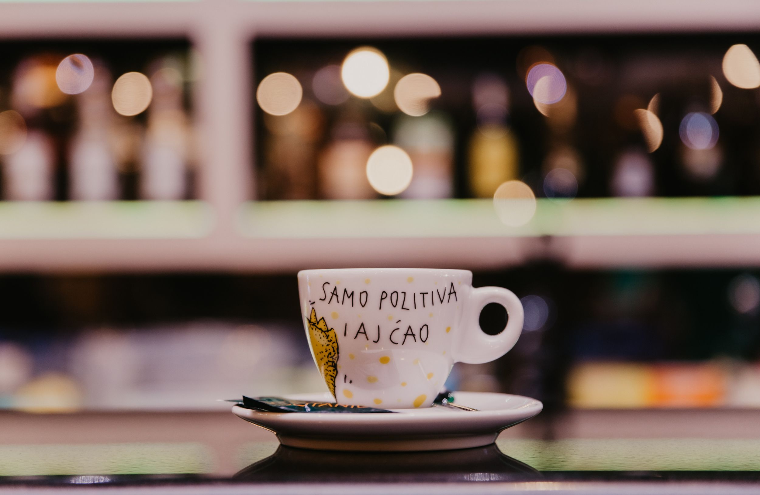 Coffee cup with 'Sans Polution' text sitting on saucer against blurred bar background.