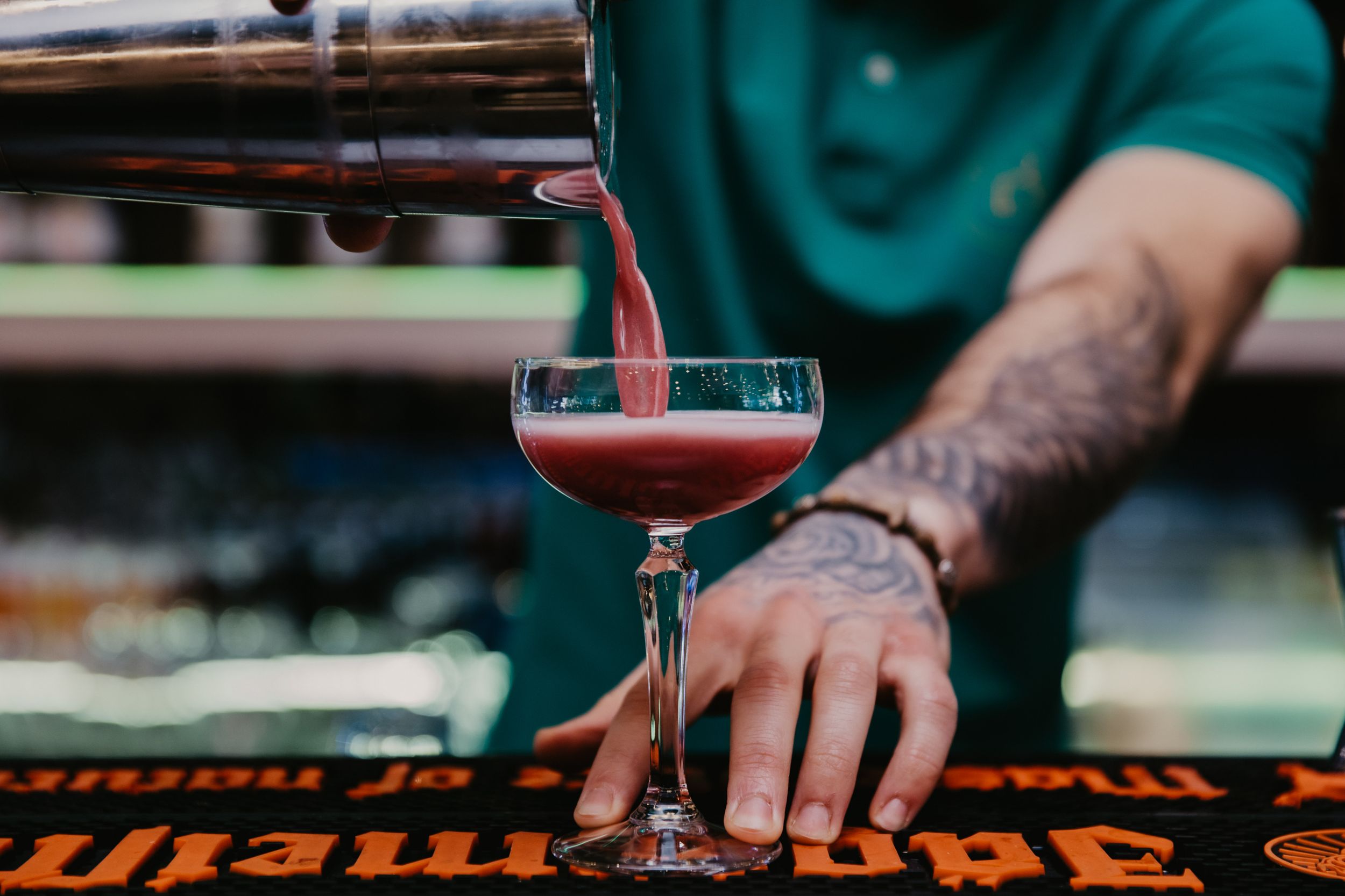 Bartender pouring red cocktail into stemmed glass at bar counter.