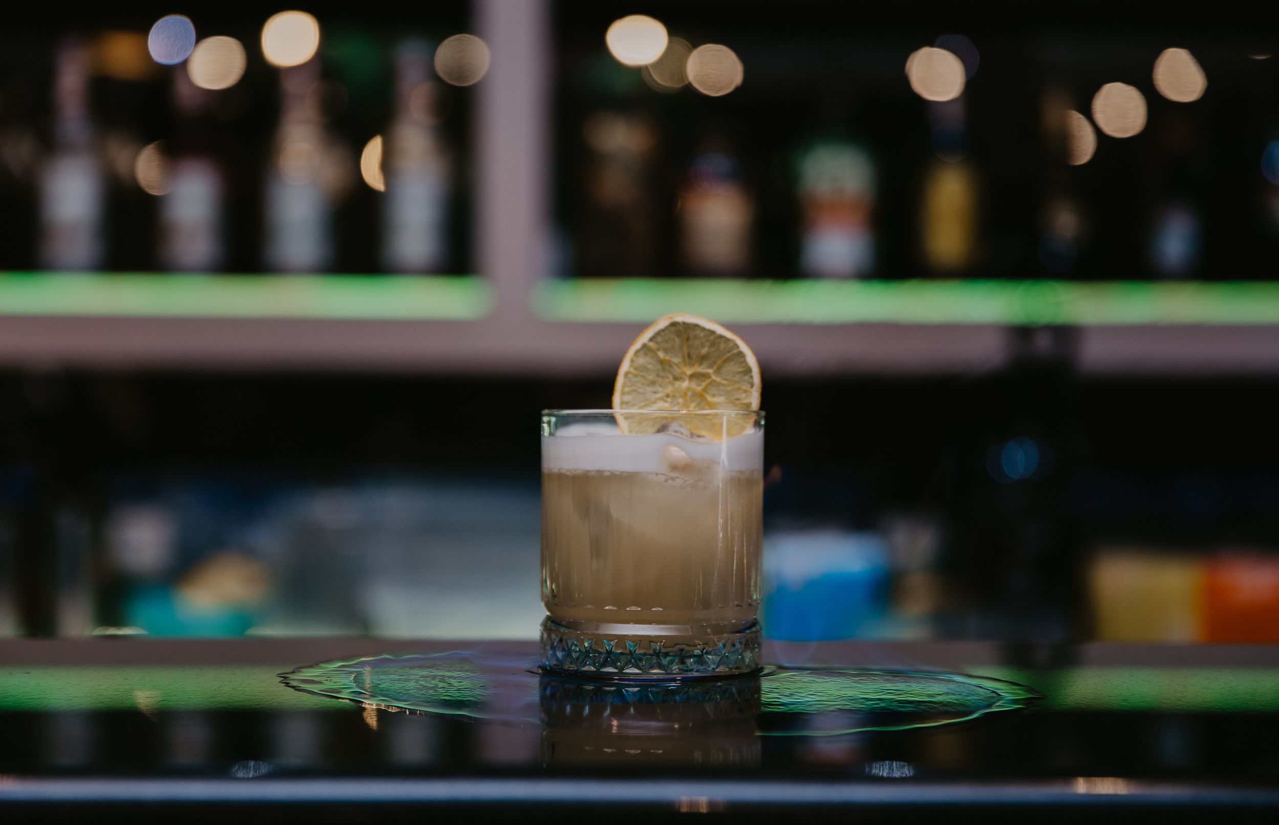 Light-colored cocktail with citrus garnish on dark bar counter with bokeh background.