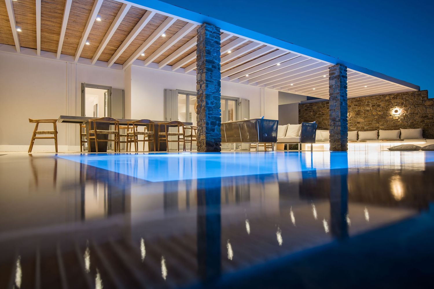 Modern villa interior with illuminated infinity pool and wooden pergola reflecting in calm waters at dusk.