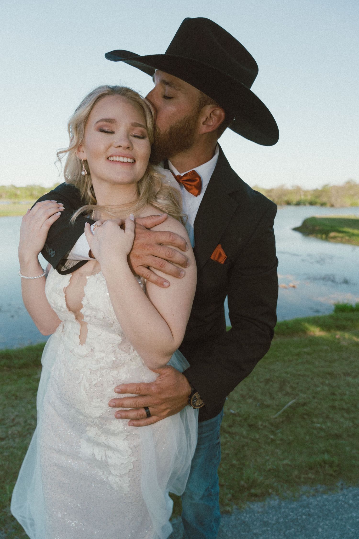 Romantic wedding photo by a lake with happy couple embracing, groom in cowboy hat and black suit.