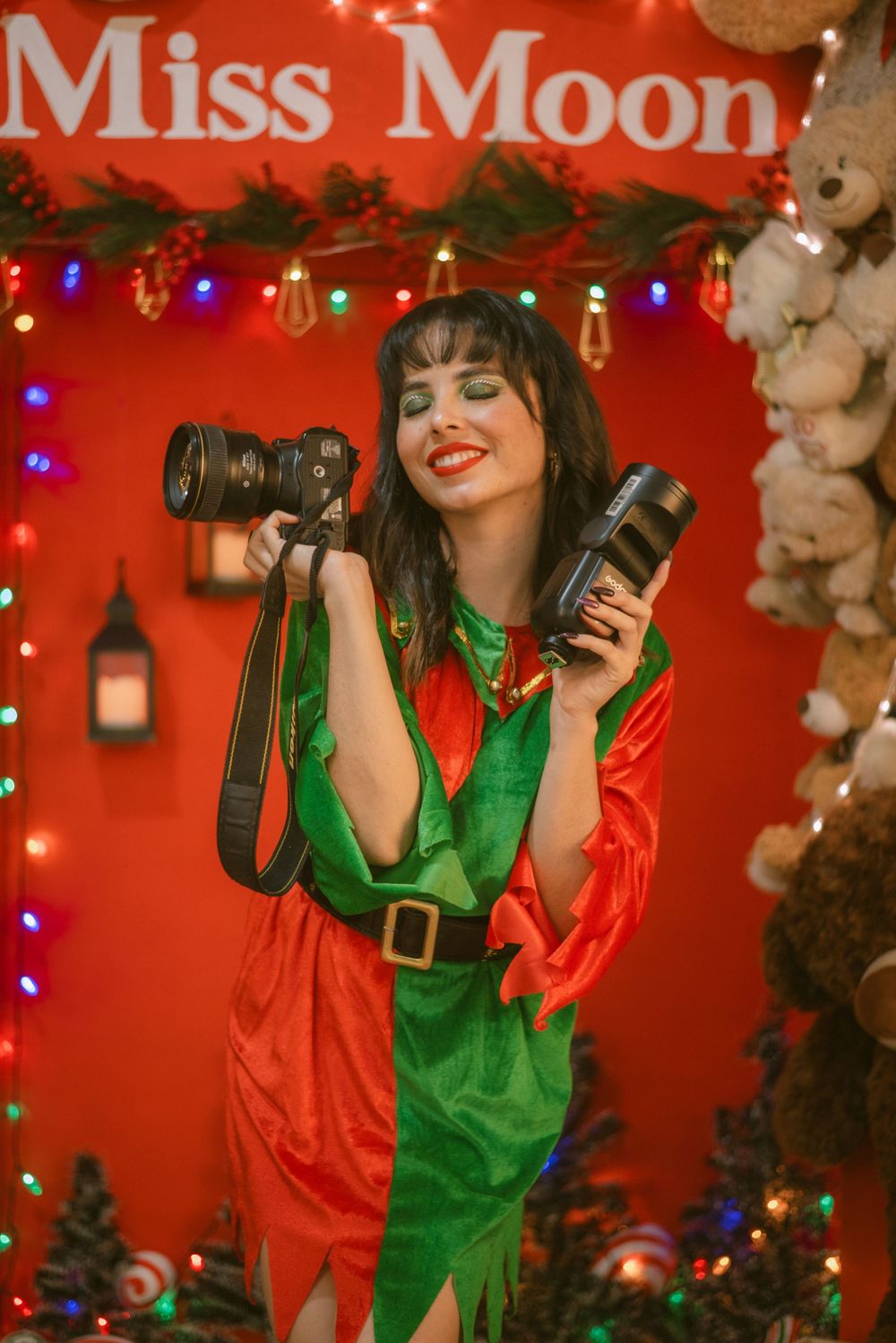 A photographer in a festive red and green elf costume holds camera equipment against a backdrop with holiday lights.
