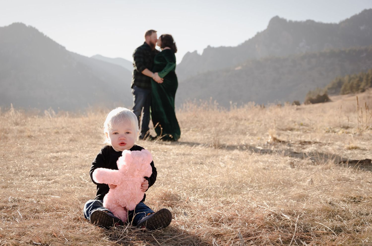 A small child sits with a pink stuffed animal while two figures embrace in the mountainous background.