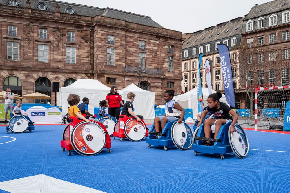 Wheelchair basketball players compete on a bright blue court during an outdoor sporting event.