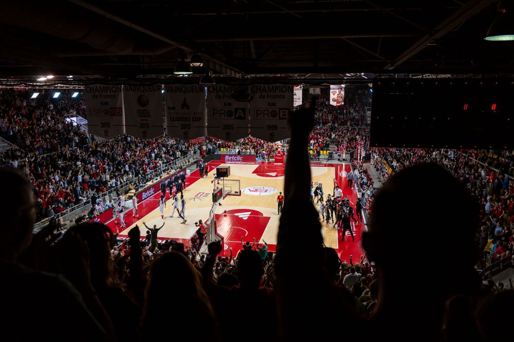 Basketball game in progress on a red court viewed from crowded stadium stands.