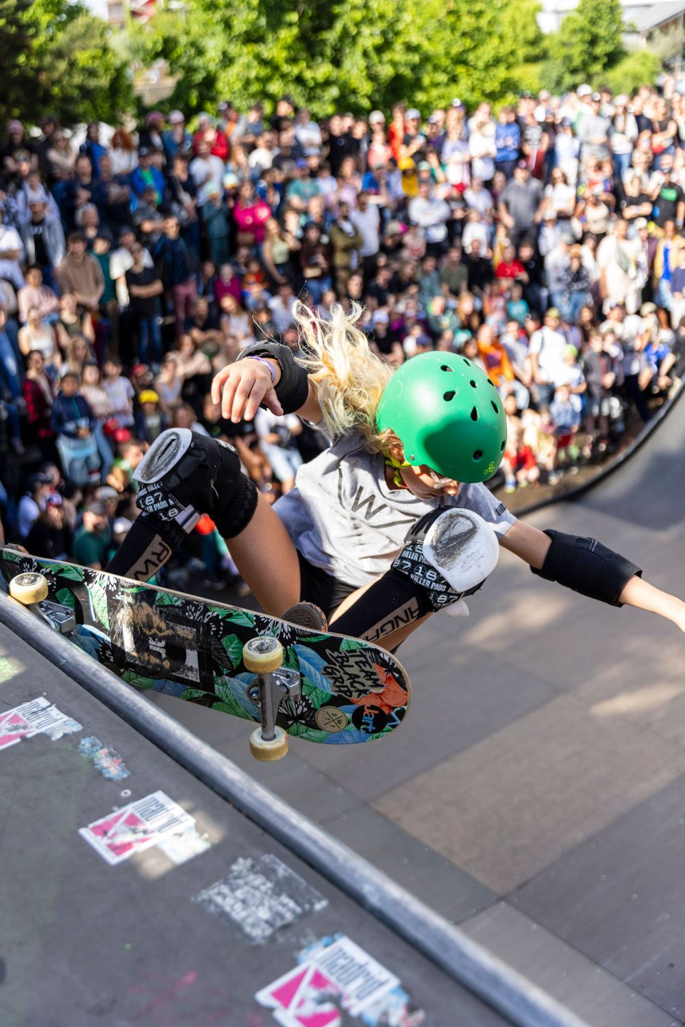Skateboarder performing aerial trick on ramp wearing protective gear with crowd watching below.