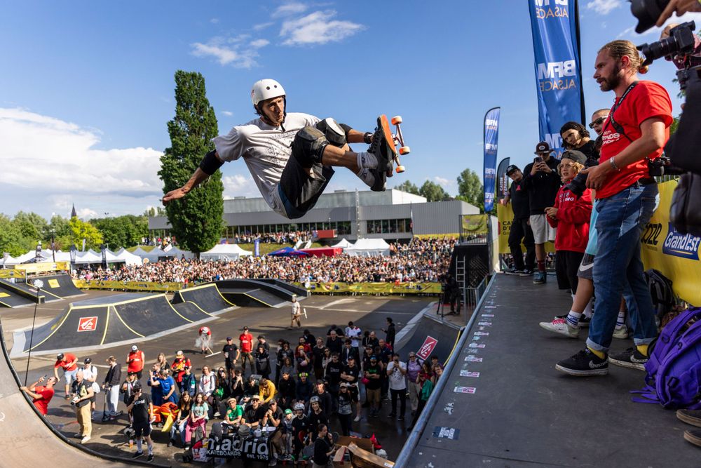 Athlete performing aerial maneuver at outdoor skate park with large crowd of spectators.
