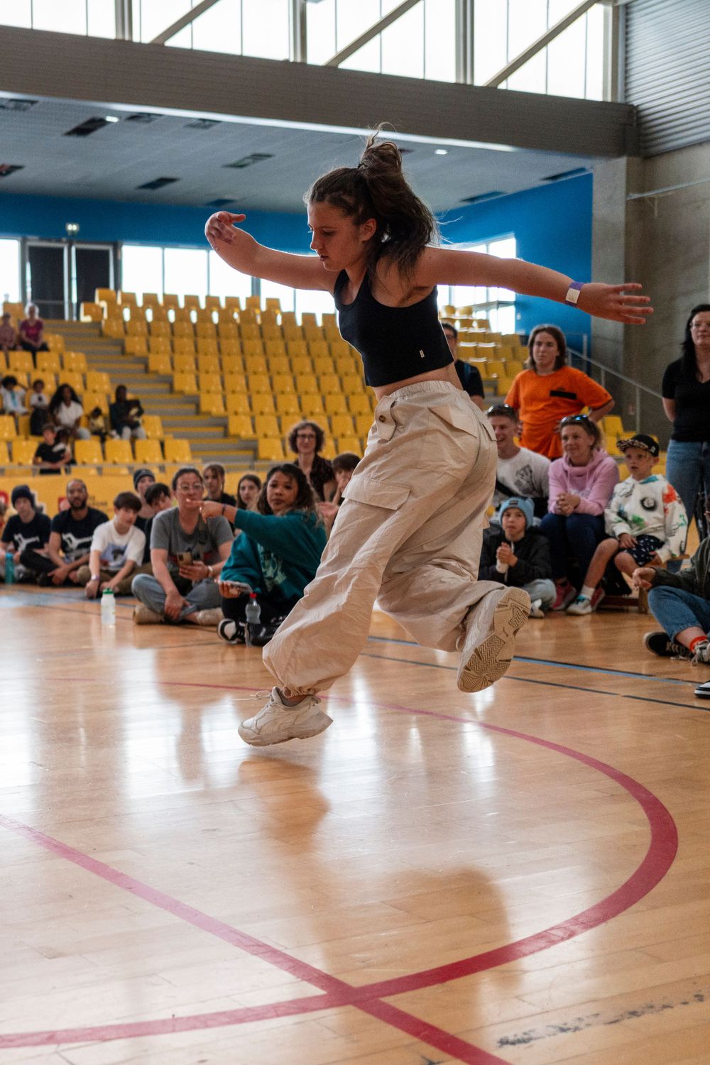 Dancer performing dynamic jump move in gymnasium with yellow seats and audience in background.