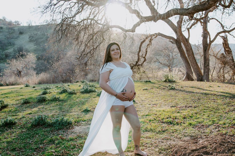 Ethereal maternity photography in a natural setting with golden sunlight filtering through trees.