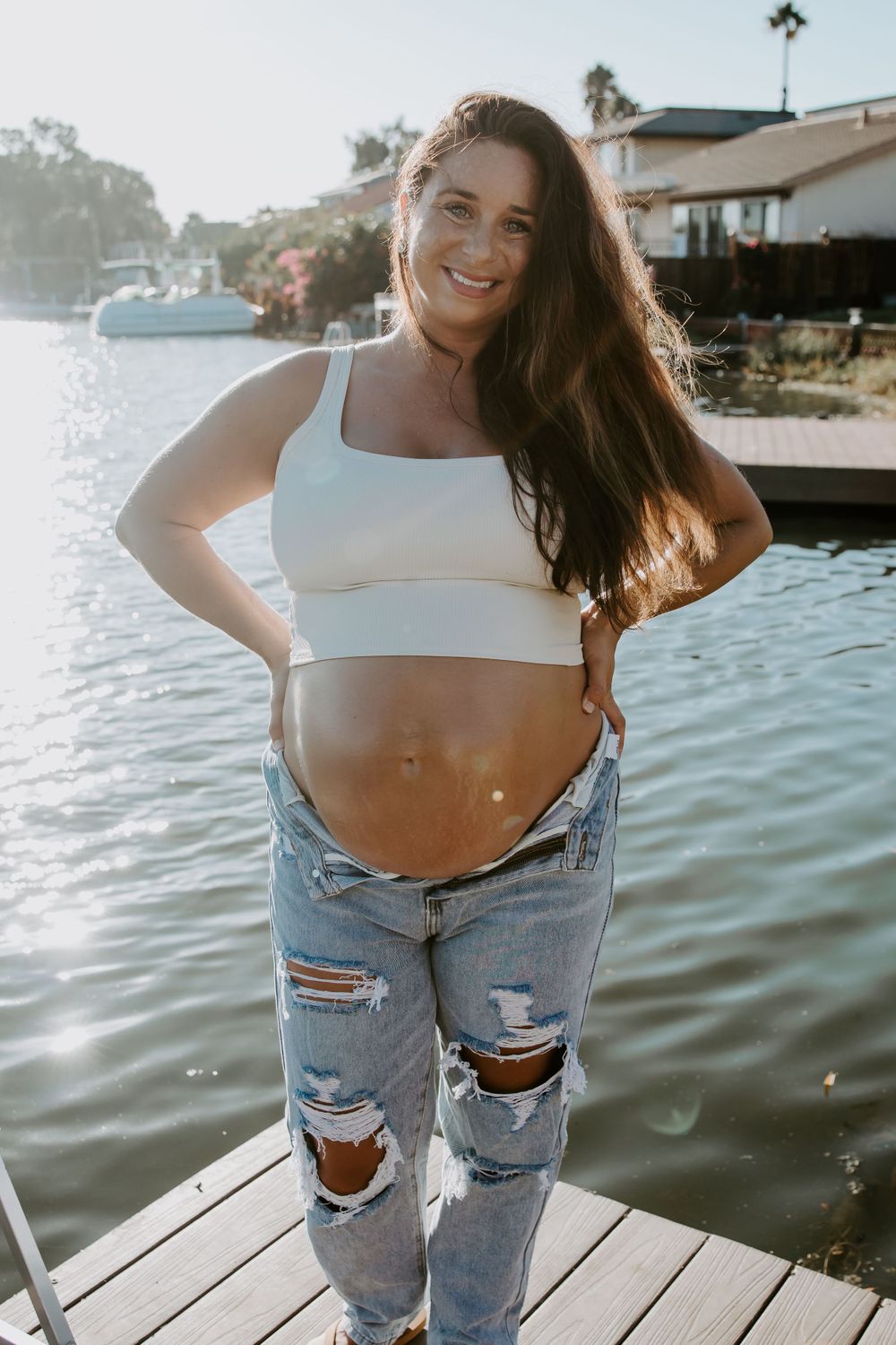 A pregnant woman in ripped jeans and white crop top poses on a dock at sunset.