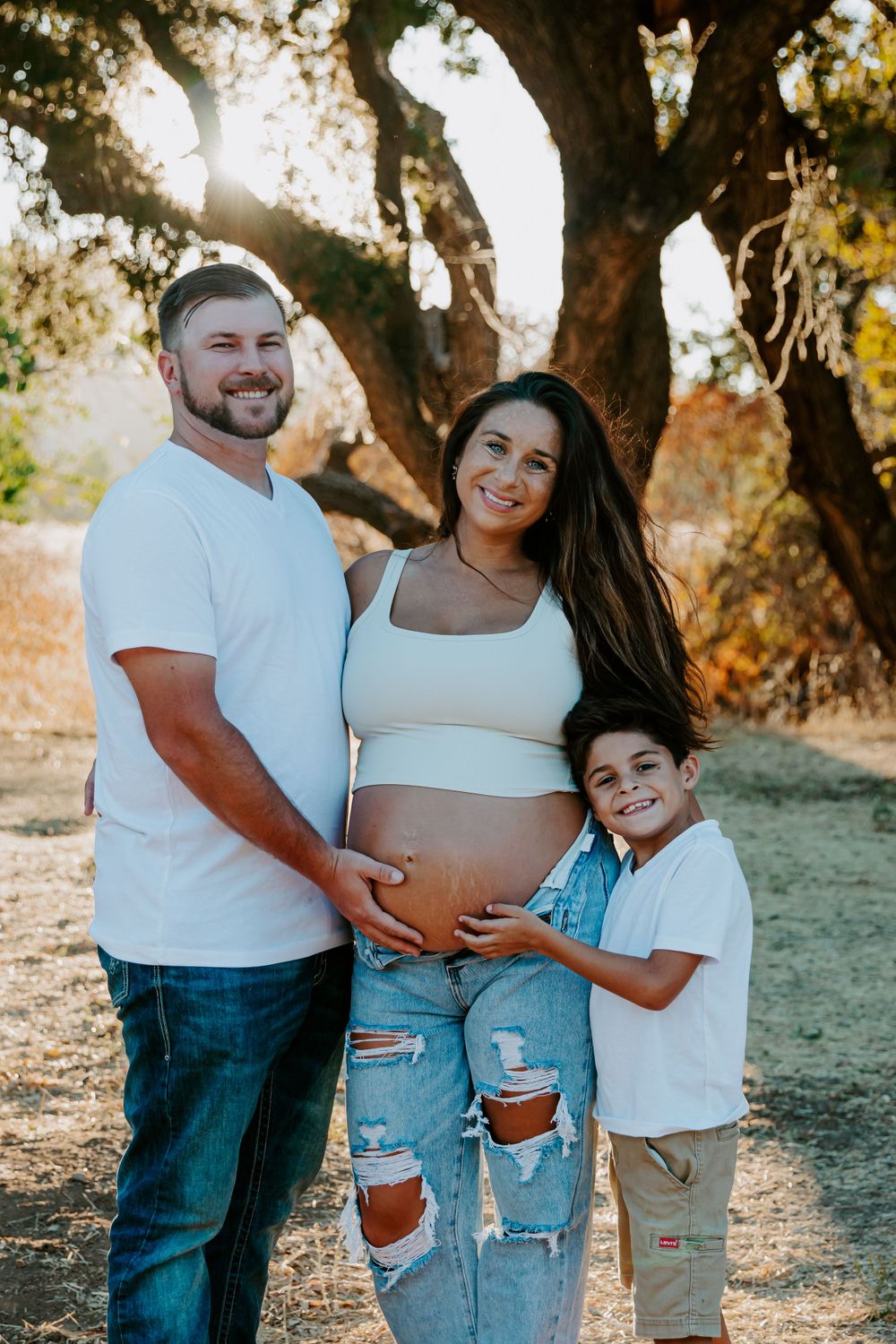 A family maternity photo with parents and child standing together under a tree at sunset.