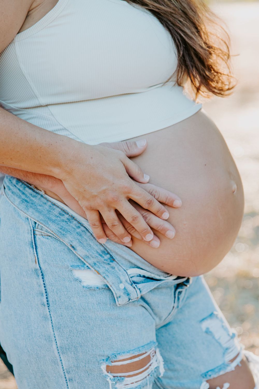Close up of pregnant belly with hands gently resting on it, wearing distressed denim.