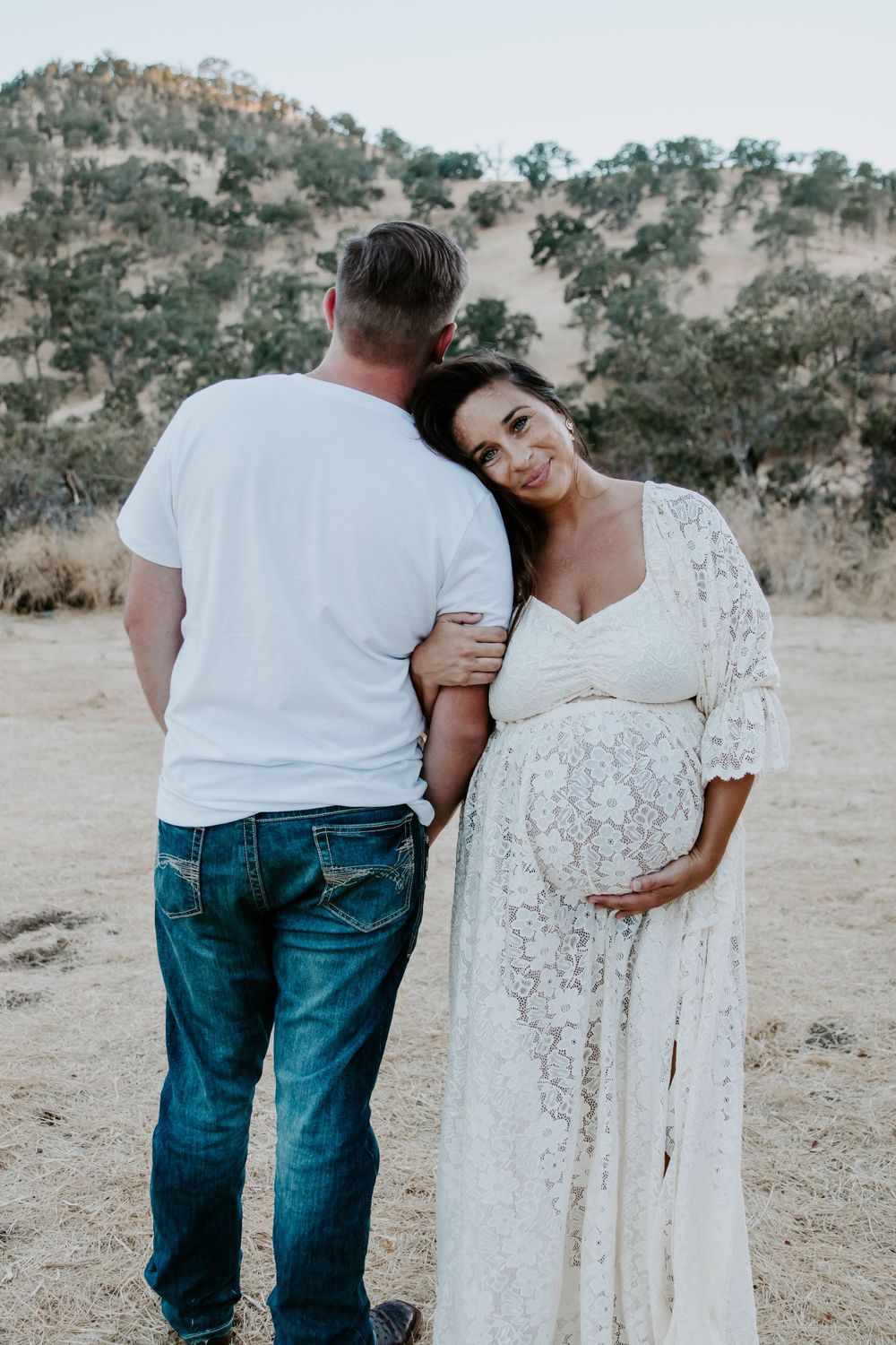 A romantic maternity portrait in a white lace dress against a mountain backdrop.